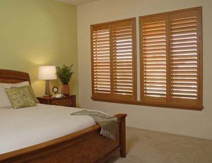 A bedroom with a queen-sized bed on the left, a nightstand with a lamp and a plant next to it, and a set of wood shutters on the right.