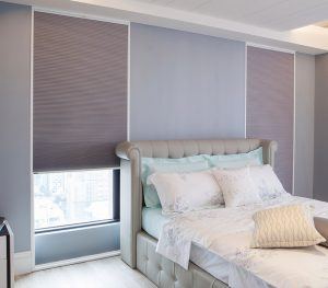 A bedroom. There is a bed in the middle of two large windows with purple roller shades.