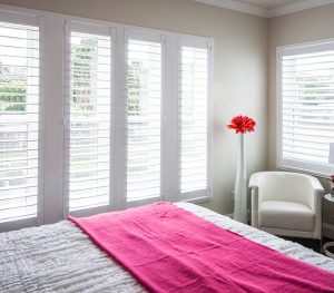 The view of a corner of a bedroom. There are lots of windows with white shutters.