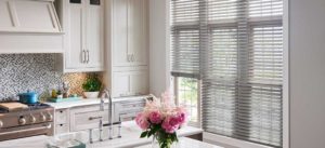 A picture of a kitchen that has a big window on the right with gray horizontal blinds