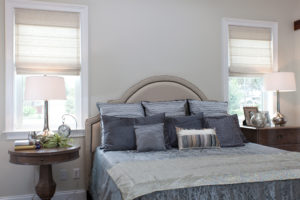 A bedroom with a queen-sized bed and two end tables on each side. Behind each end table is a window with Roman shades half-rolled up.