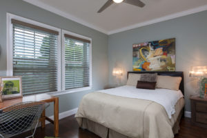 A bedroom with a queen-sized bed and two double-hung windows on the far wall. There are brown horizontal blinds on the windows.