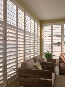 The corner of a living room with windows lining the walls. The windows have white shutters.