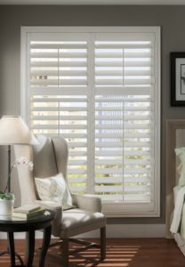 A bedroom with a large white window shutters. A beige wingback chair with a lamp sits in front of the window.