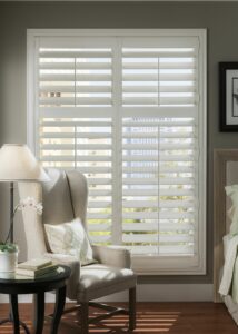 A corner of a bedroom with a chair and side table beside a large window with white shutters