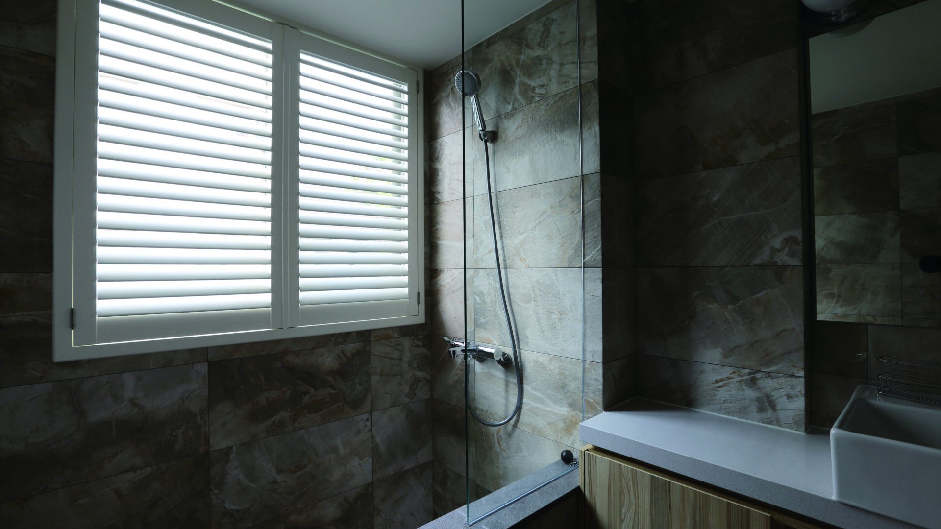 White Woodlore Plus plantation shutters installed on a bathroom window next to a glass shower enclosure with natural stone tile