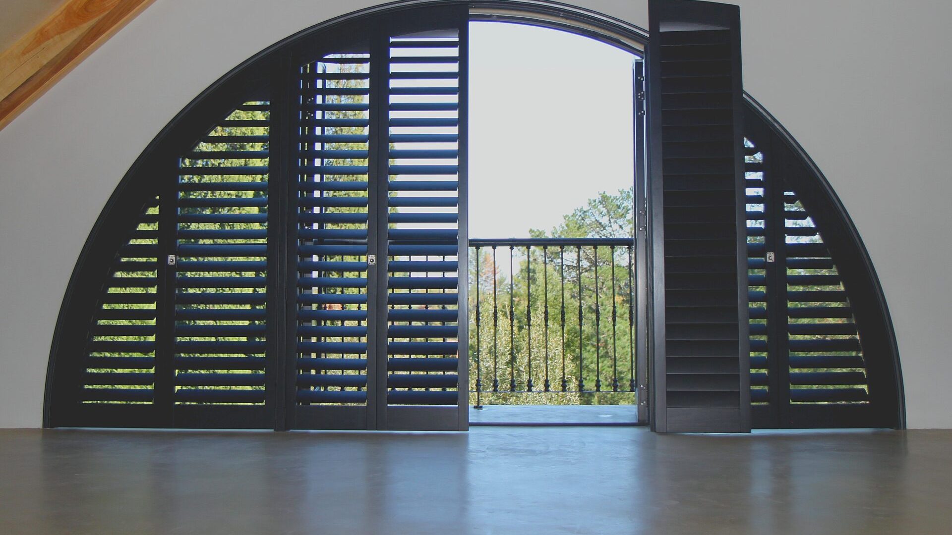 Dark-stained specialty shape plantation shutters custom-fitted to a large semi-circle arched doorway opening to a balcony with tree views