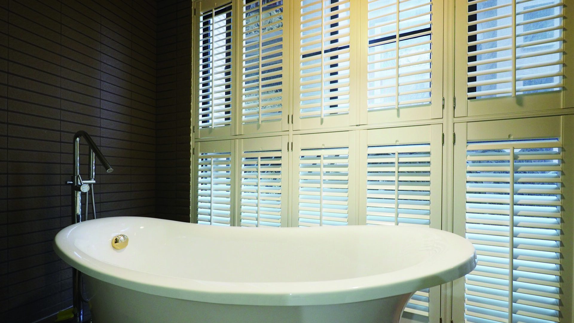Floor-to-ceiling white Woodlore Plus plantation shutters behind a freestanding bathtub in an elegant spa-style bathroom with a dark teal accent wall