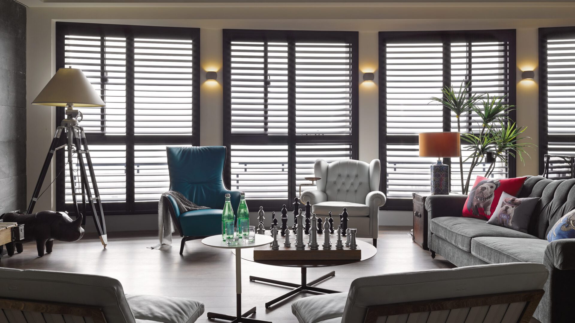 Dark-stained Normandy hardwood plantation shutters spanning multiple tall windows in a sophisticated living room with designer furniture and ambient lighting