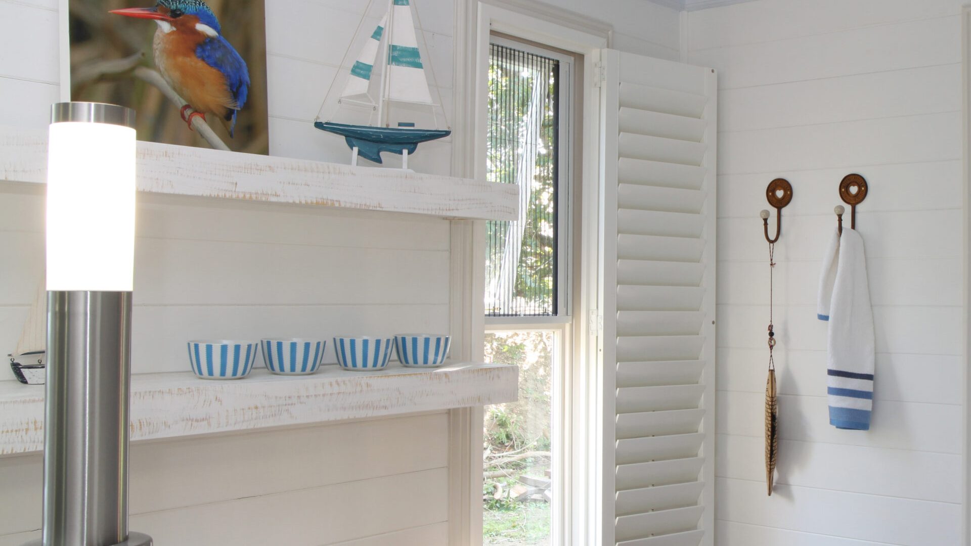 White Woodlore plantation shutters on a small window in a bright coastal cottage interior with nautical decor