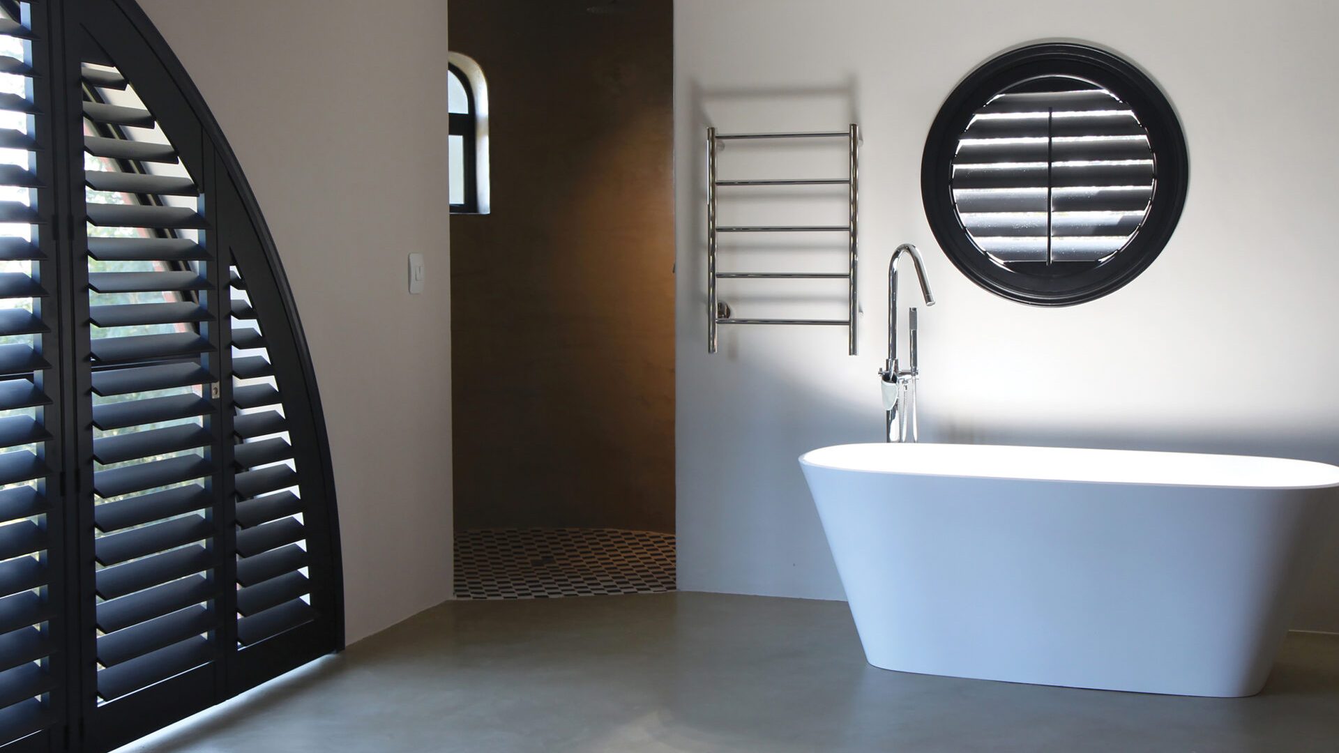 Modern bathroom featuring dark-stained arched and round porthole specialty shape plantation shutters with a freestanding bathtub and heated towel rack